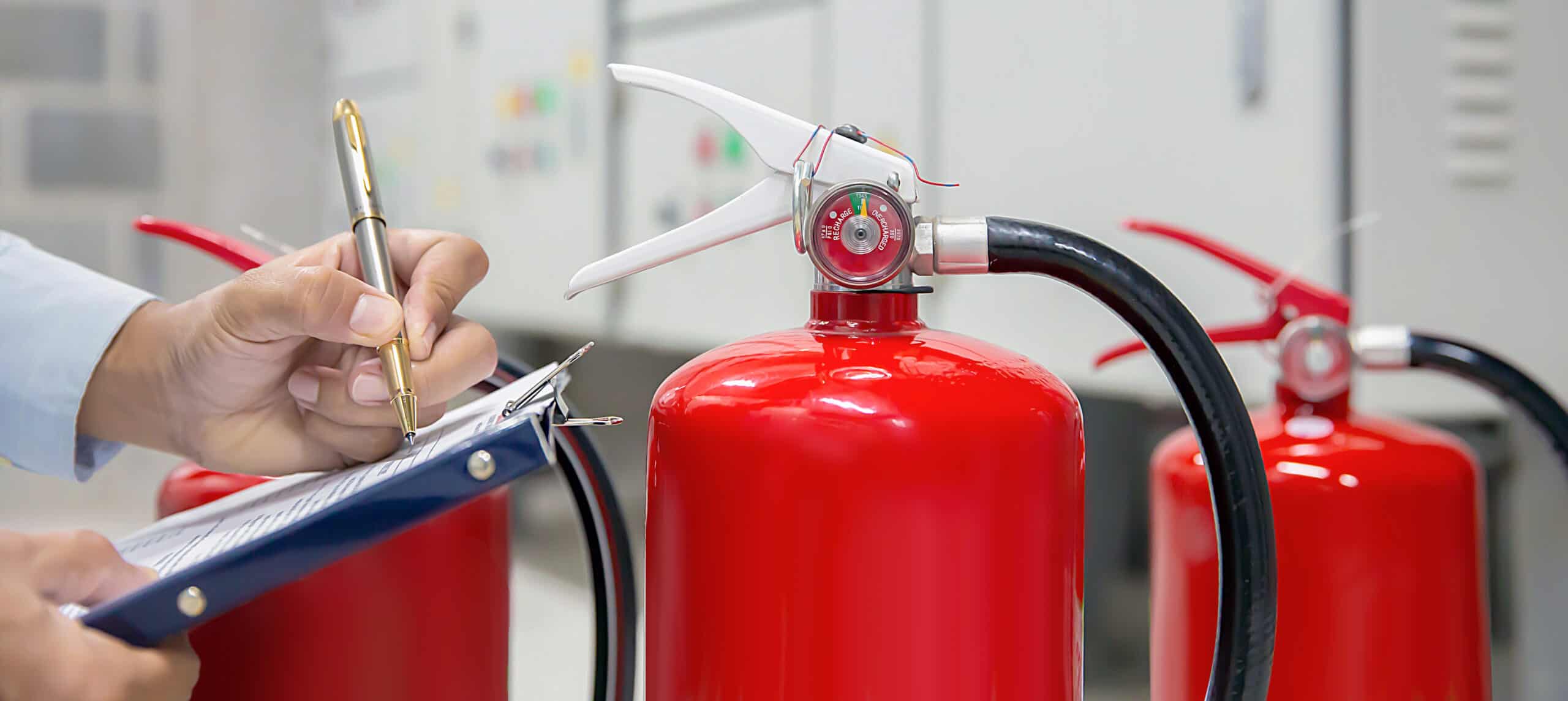 A person writes on a checklist attached to a clipboard while inspecting bright red fire extinguishers positioned in front of electrical control panels.