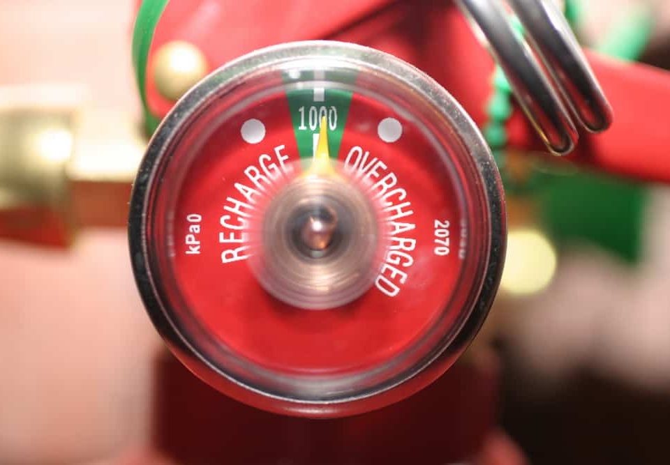 A close-up view of a fire extinguisher pressure gauge showing markings for “Recharge” and “Overcharged,” with the needle pointing to the green zone indicating proper pressure.