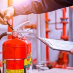 A person holds a clipboard with a checklist and uses a pen to inspect a red fire extinguisher in an indoor area with visible pipes and valves in the background.