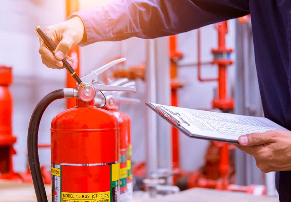 A person holds a clipboard with a checklist and uses a pen to inspect a red fire extinguisher in an indoor area with visible pipes and valves in the background.