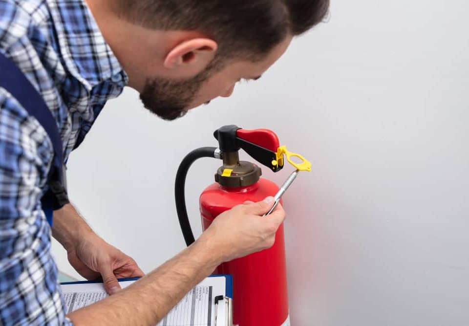 A person inspects a red fire extinguisher mounted on a wall, holding a pen and clipboard while checking the safety seal and pin.