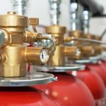 A row of red fire suppression cylinders with brass valves and pressure gauges connected to metal piping, arranged in an indoor setting.
