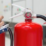 A person holding a pen marks a checklist on a clipboard while inspecting bright red fire extinguishers arranged in front of electrical control panels.