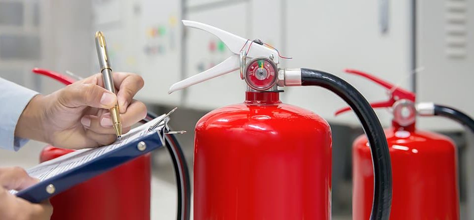 A person holding a pen marks a checklist on a clipboard while inspecting bright red fire extinguishers arranged in front of electrical control panels.