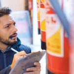 A person holds a clipboard and pen while inspecting a row of red fire extinguishers mounted vertically on a wall.