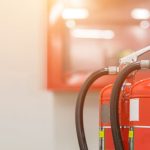 Three red fire extinguishers with black hoses are positioned in the foreground, highlighting Fire Extinguishers Sales & Service in Long Island, with a blurred indoor background featuring a glass-fronted emergency equipment cabinet.