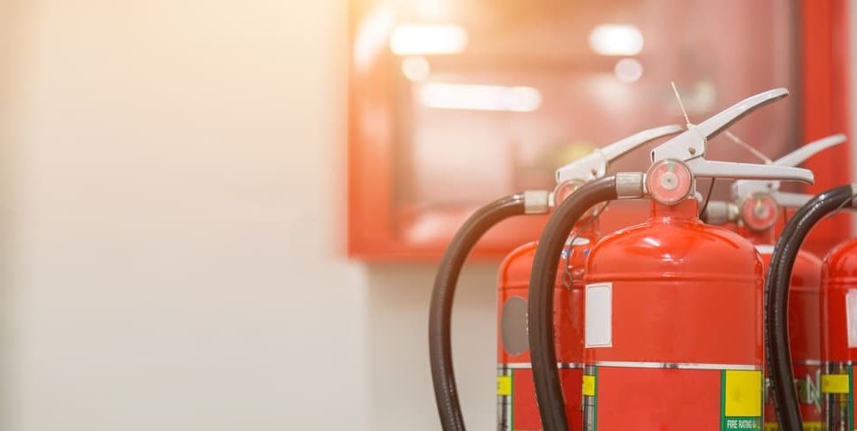 Three red fire extinguishers with black hoses are positioned in the foreground, highlighting Fire Extinguishers Sales & Service in Long Island, with a blurred indoor background featuring a glass-fronted emergency equipment cabinet.
