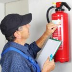 A man in a blue shirt and cap inspects a red fire extinguisher mounted on a white wall, holding a clipboard and pen as he examines the label—showcasing Fire Extinguishers Sales & Service in Long Island.