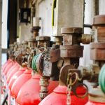 A row of red fire extinguishers with metal valves and hoses lines a wall on a ship, the ocean visible behind, showcasing Fire Extinguishers Sales & Service in Long Island for marine or industrial safety.