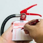 A hand holds a fire extinguisher while another marks an inspection record tag with a pen. The tag lists months and years for maintenance checks, ensuring compliance with Fire Extinguishers Sales & Service in Long Island standards.