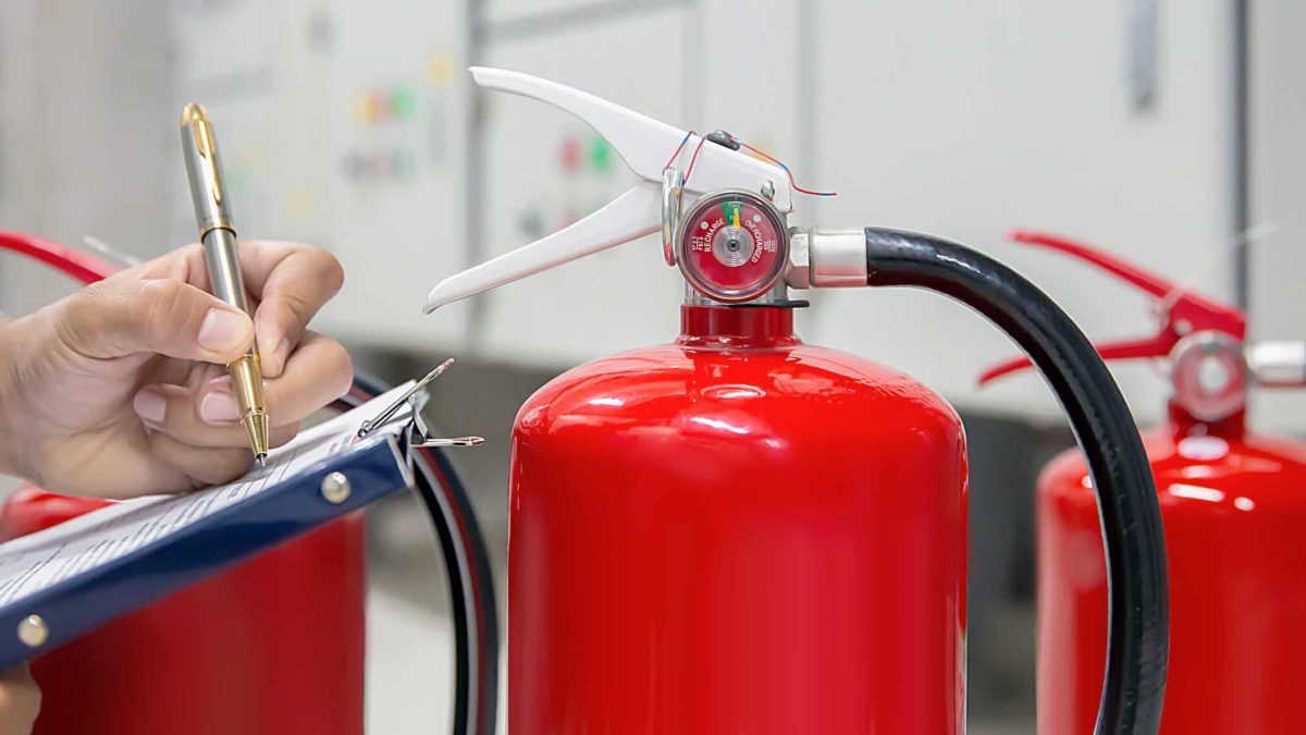 A person inspects a red fire extinguisher and takes notes on a clipboard, with two others visible in the background—highlighting Fire Extinguishers Sales & Service in Long Island within an indoor facility or control room.