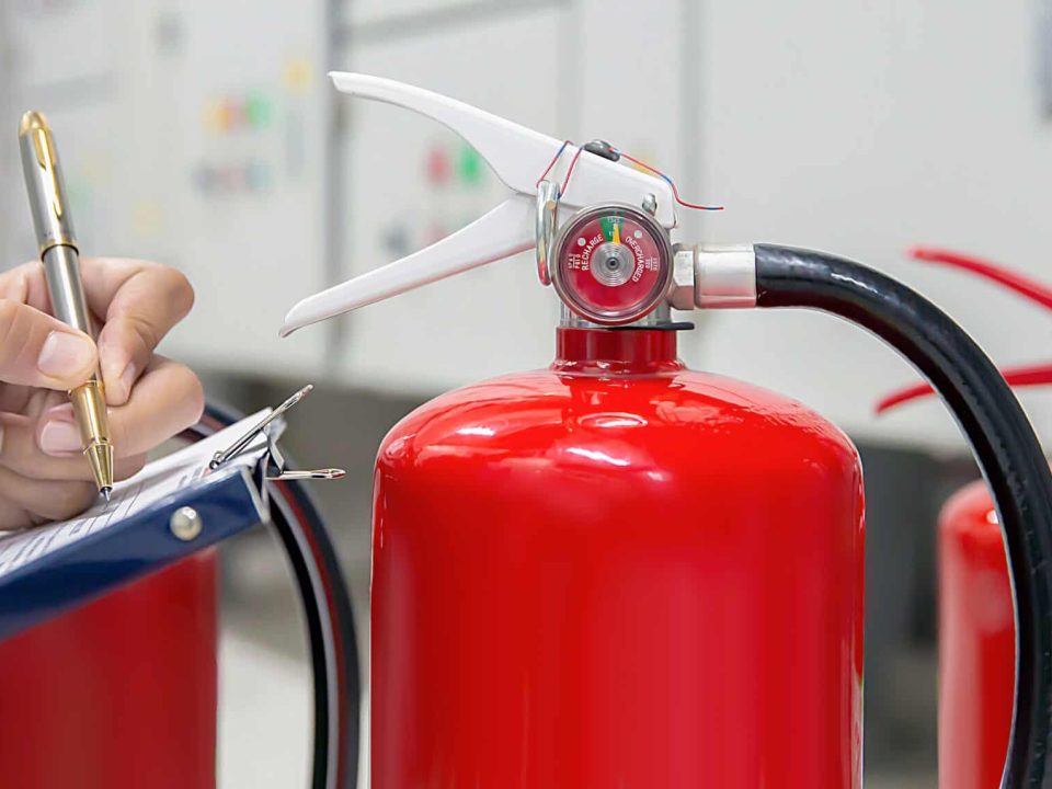 A person inspects a red fire extinguisher and takes notes on a clipboard, with two others visible in the background—highlighting Fire Extinguishers Sales & Service in Long Island within an indoor facility or control room.
