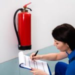A woman in a blue uniform kneels next to a wall-mounted fire extinguisher, writing on a clipboard during an inspection for Fire Extinguishers Sales & Service in Long Island.