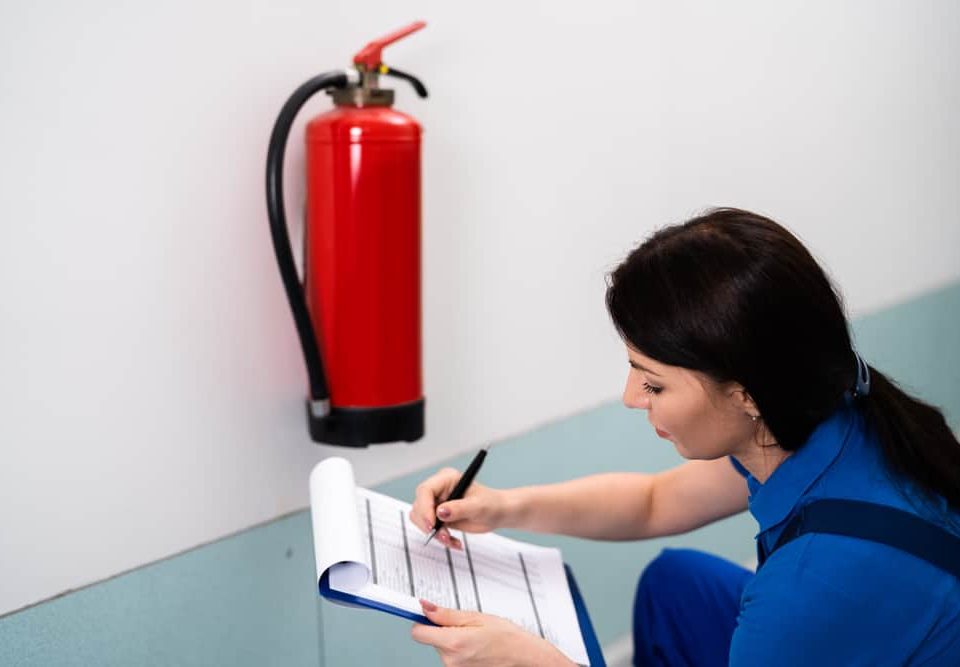 A woman in a blue uniform kneels next to a wall-mounted fire extinguisher, writing on a clipboard during an inspection for Fire Extinguishers Sales & Service in Long Island.