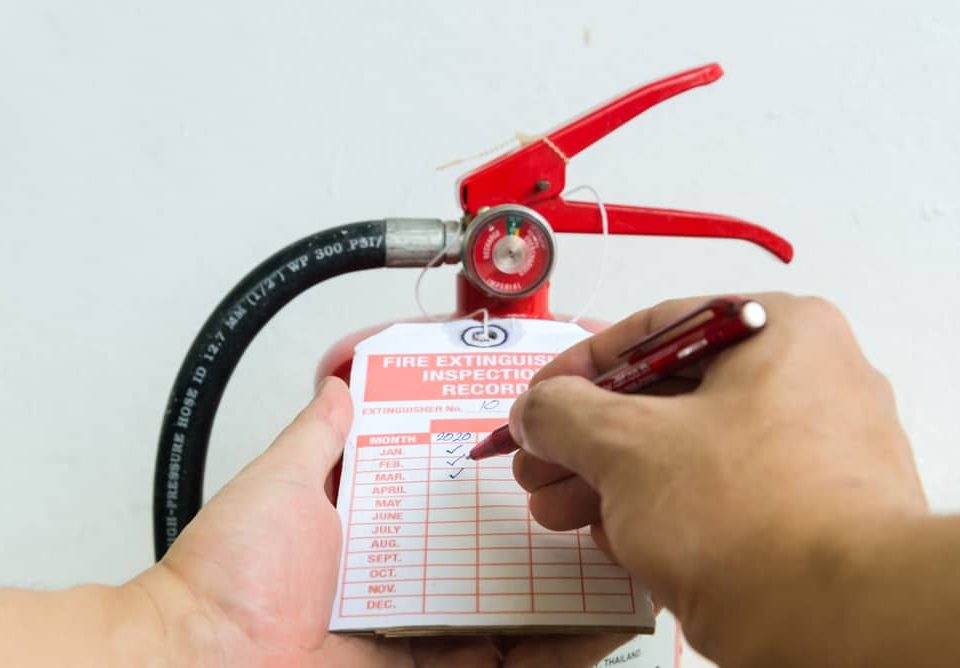 A hand holds a fire extinguisher while another marks an inspection record tag with a pen. The tag lists months and years for maintenance checks, ensuring compliance with Fire Extinguishers Sales & Service in Long Island standards.