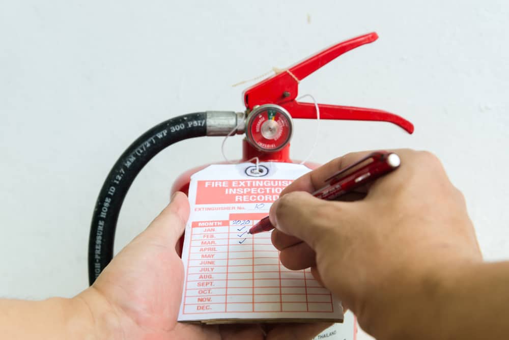 A hand holds a fire extinguisher while another marks an inspection record tag with a pen. The tag lists months and years for maintenance checks, ensuring compliance with Fire Extinguishers Sales & Service in Long Island standards.
