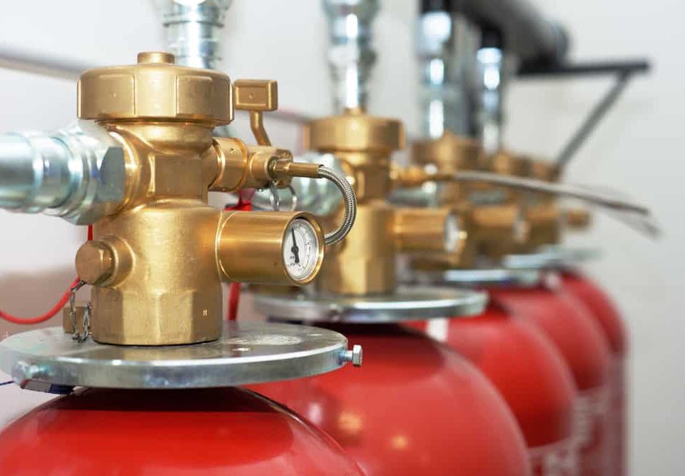 Close-up view of several red fire suppression system cylinders with brass valves and pressure gauges, arranged in a row and connected by metal pipes—ideal for those seeking Fire Extinguishers Sales & Service in Long Island.