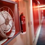 A close-up of a fire hose coiled in a wall-mounted case, with a fire extinguisher and the brightly lit red hallway of a Fire Extinguishers Sales & Service in Long Island location in the background.