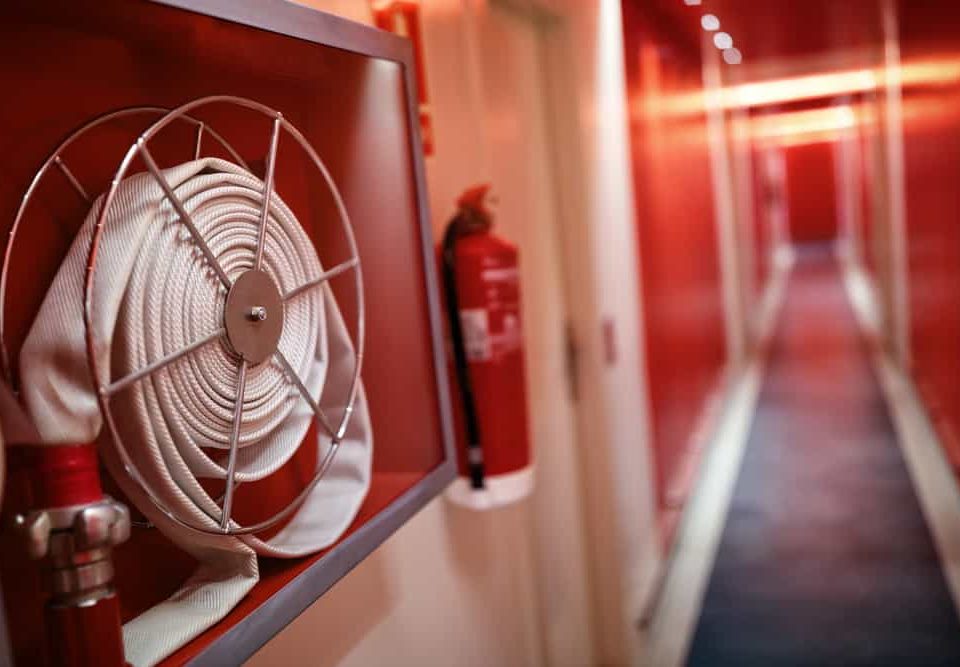 A close-up of a fire hose coiled in a wall-mounted case, with a fire extinguisher and the brightly lit red hallway of a Fire Extinguishers Sales & Service in Long Island location in the background.