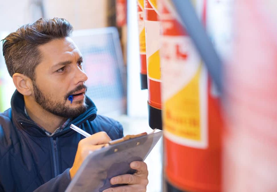 A man with a pen in his mouth holds a clipboard and inspects a row of red fire extinguishers, appearing focused and thoughtful, representing Fire Extinguishers Sales & Service in Long Island.